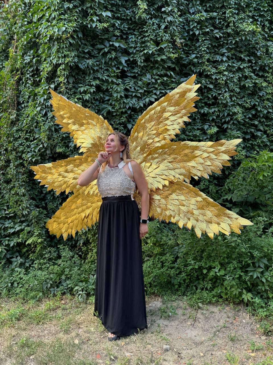 May include: A woman wearing a gold sequined top and a long black skirt, posing in front of a wall of green foliage. Large, golden, feathered wings are attached to her back, creating a striking visual effect. The wings are the focal point.