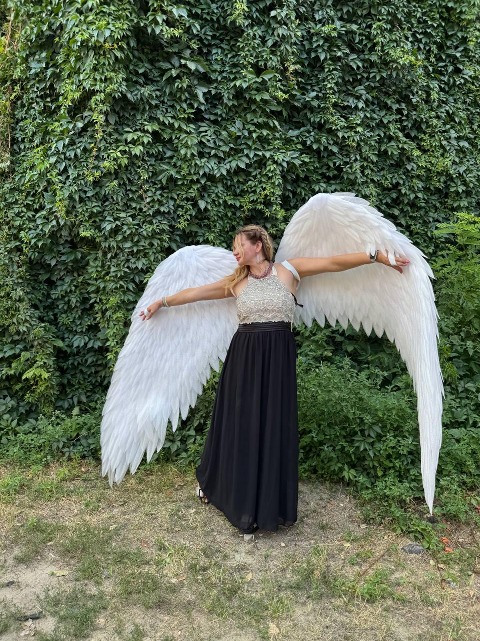May include: A person wearing large white feathered angel wings, a silver and white top, and a long black skirt, standing in front of a green leafy background. The wings are spread wide.