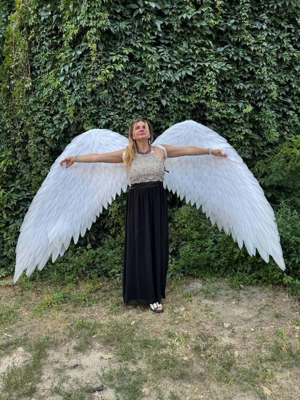 May include: A person wearing a black dress and a beaded top, with large white feathered angel wings spread wide. The background is a wall of green foliage, with grass in the foreground.