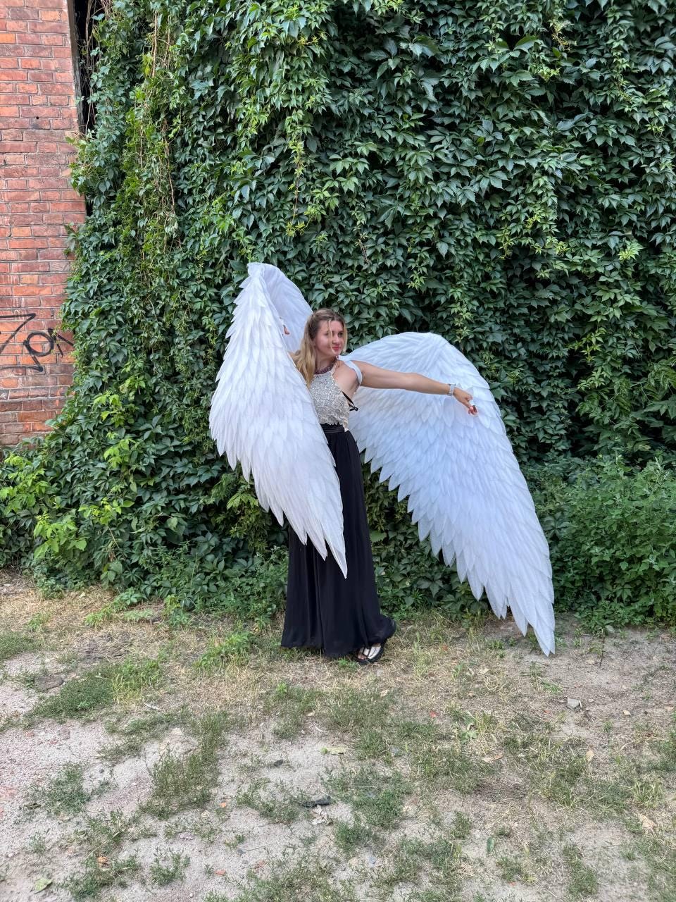 May include: A person wearing large, white angel wings, standing in front of a wall of green foliage. The wings are detailed with layered feathers. The person is wearing a black dress and a silver top.
