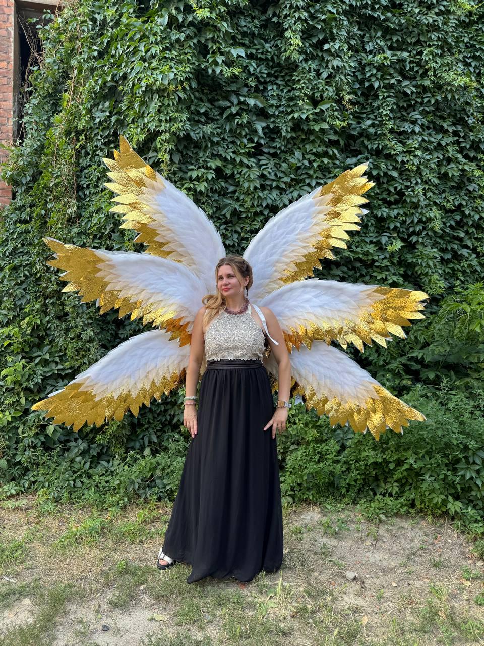 May include: A woman wearing large angel wings with white and gold feathers, standing in front of a wall of green foliage. She is wearing a silver top and a long black skirt. The wings are the focal point of the image.