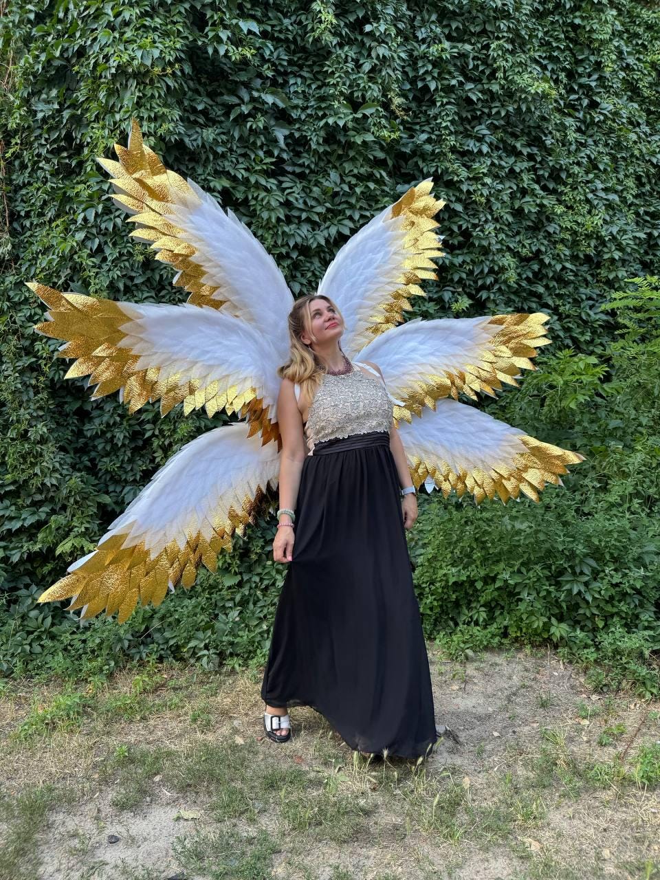 May include: A woman wearing large white and gold angel wings, a sequined top, and a long black dress. The wings have white feathers with gold glitter accents. The background is a lush green wall of foliage.