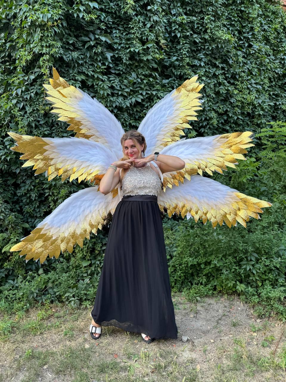 May include: Large, white and gold angel wings with a woman wearing a black skirt and a silver top. The wings have white feathers with gold tips. The woman is smiling and posing in front of a green leafy background.