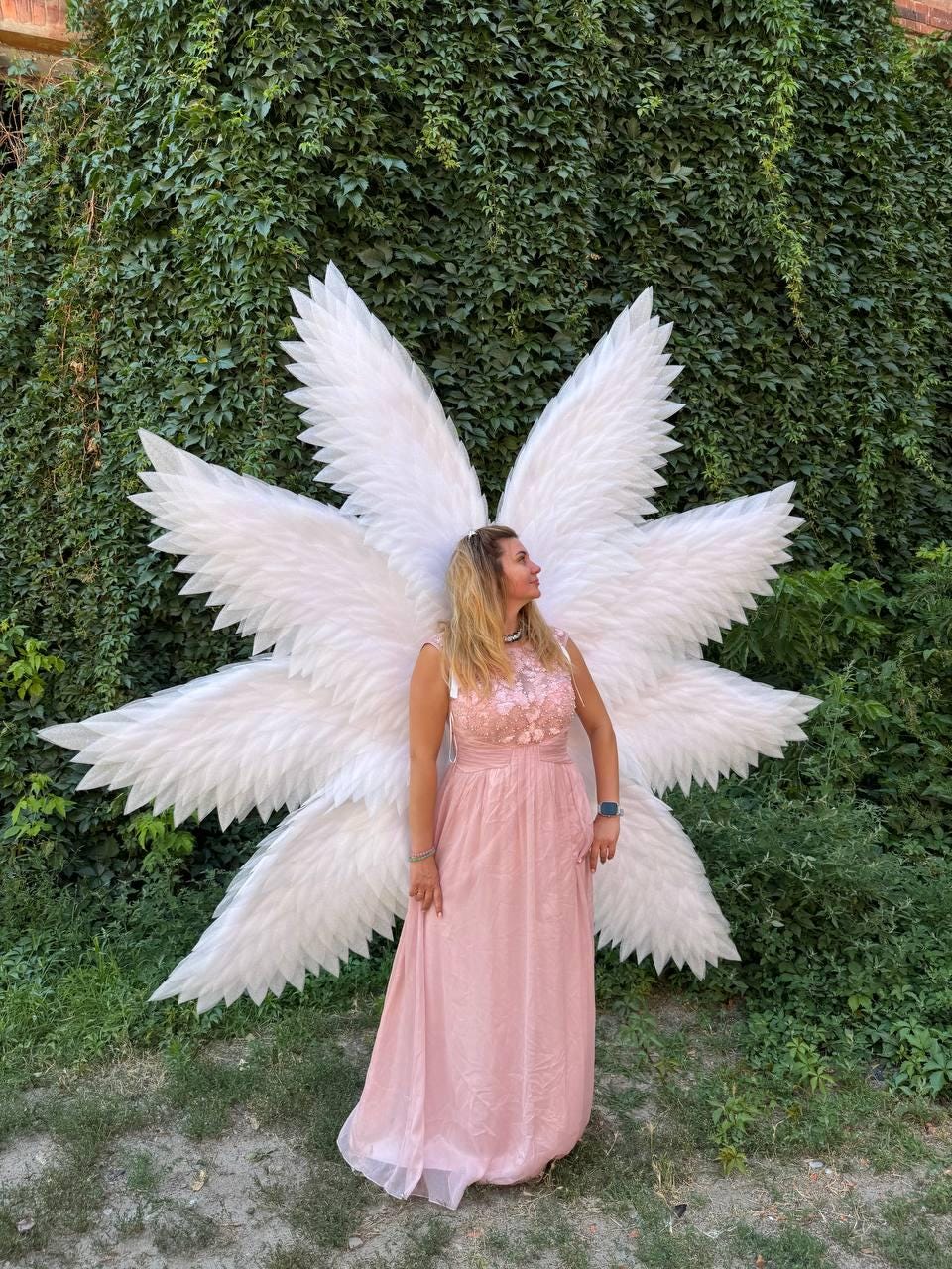 May include: A person wearing a light pink dress and large white angel wings stands in front of a green wall of foliage. The wings are large and feathered, with a starburst design. The dress has a floral bodice.