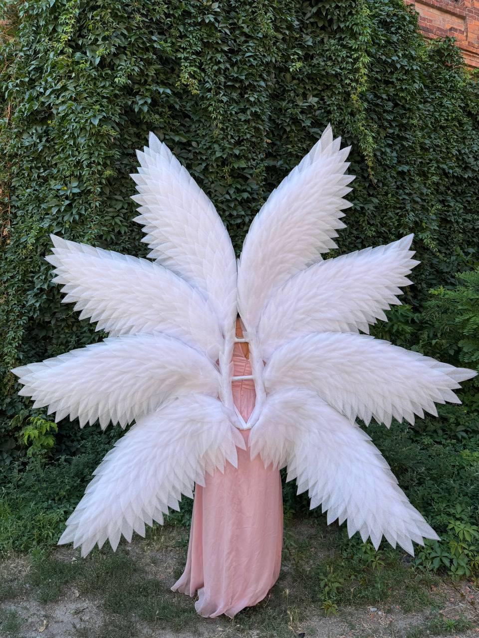 May include: A person wearing large, white angel wings with multiple layers of feathers. The wings are spread wide, and the person is wearing a long, flowing pink dress. The background is a lush green wall of foliage.