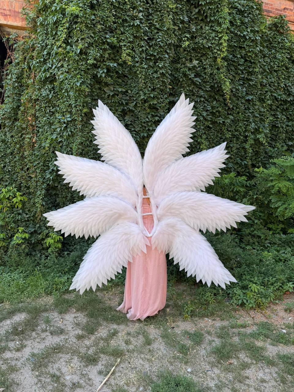 May include: A person wearing a pink dress and large white feathered wings. The wings are arranged in a circular pattern, with each wing section radiating outwards. The background features lush green foliage and a building.