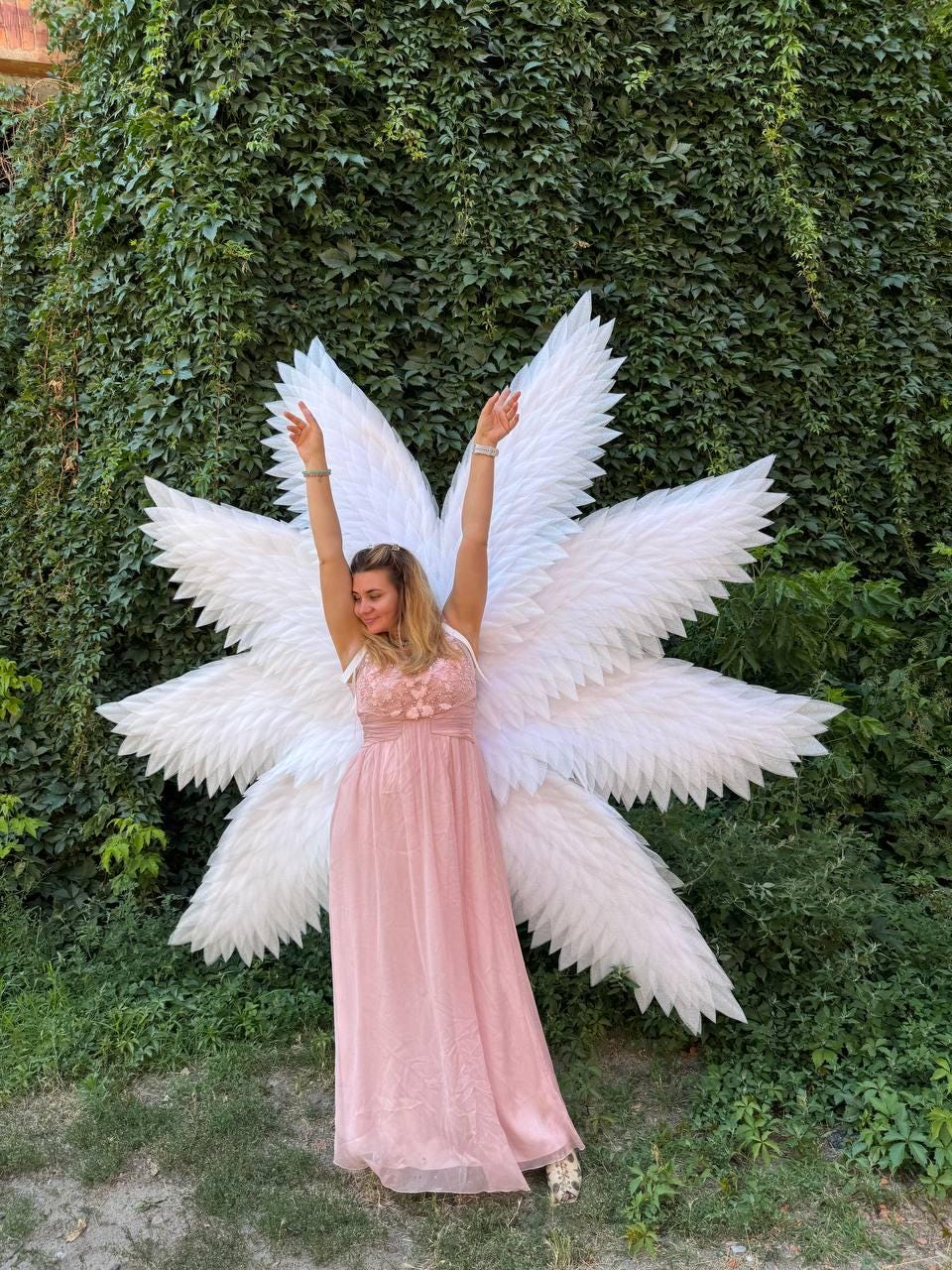 May include: A woman wearing a pink dress and large white angel wings stands with arms raised. The wings are made of layered white feathers. The background is a wall of green foliage.