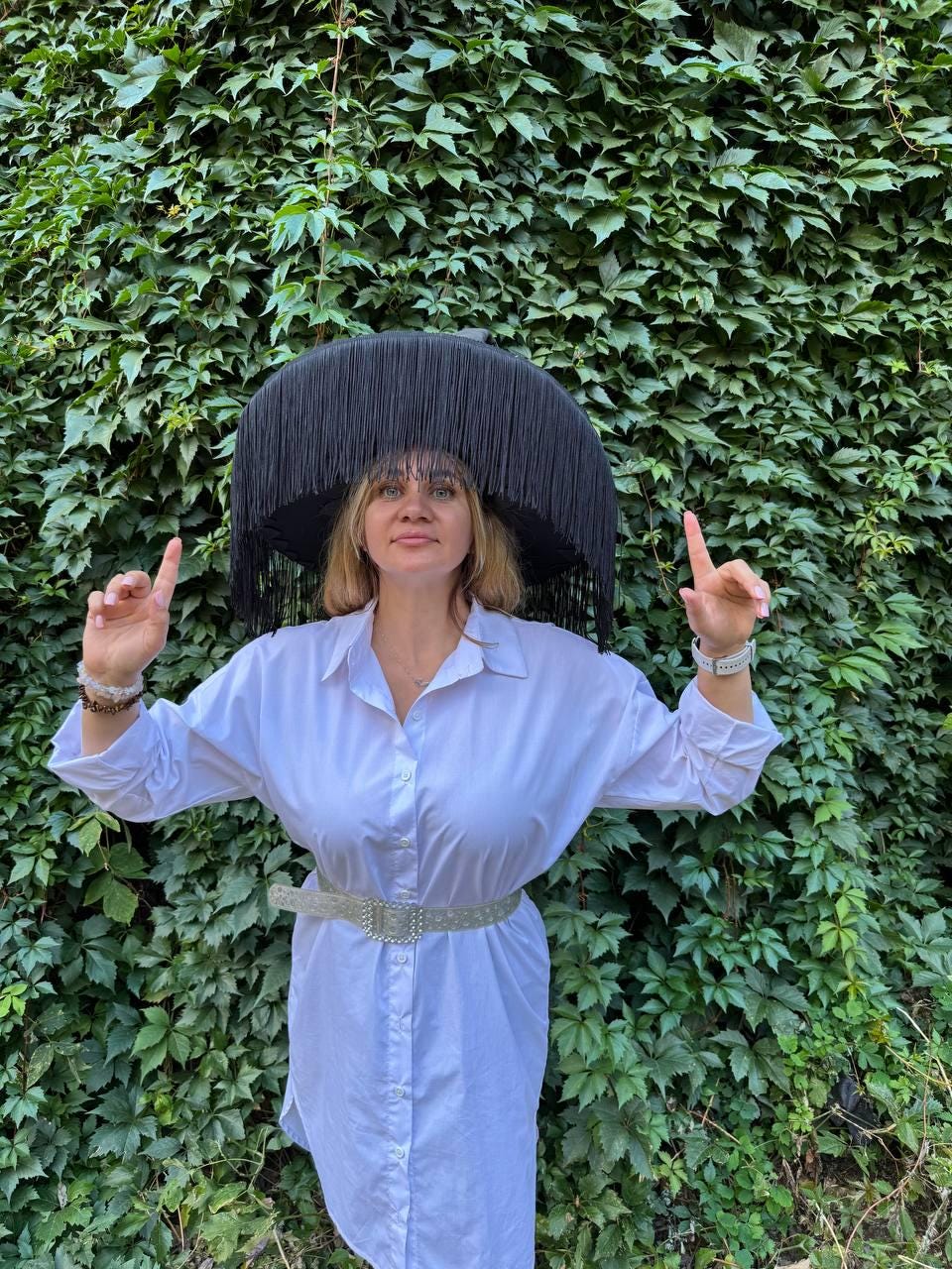 May include: A black fringed hat with a wide brim. The hat is worn with a white button-down shirt and a silver belt. The background is a wall of green leaves.