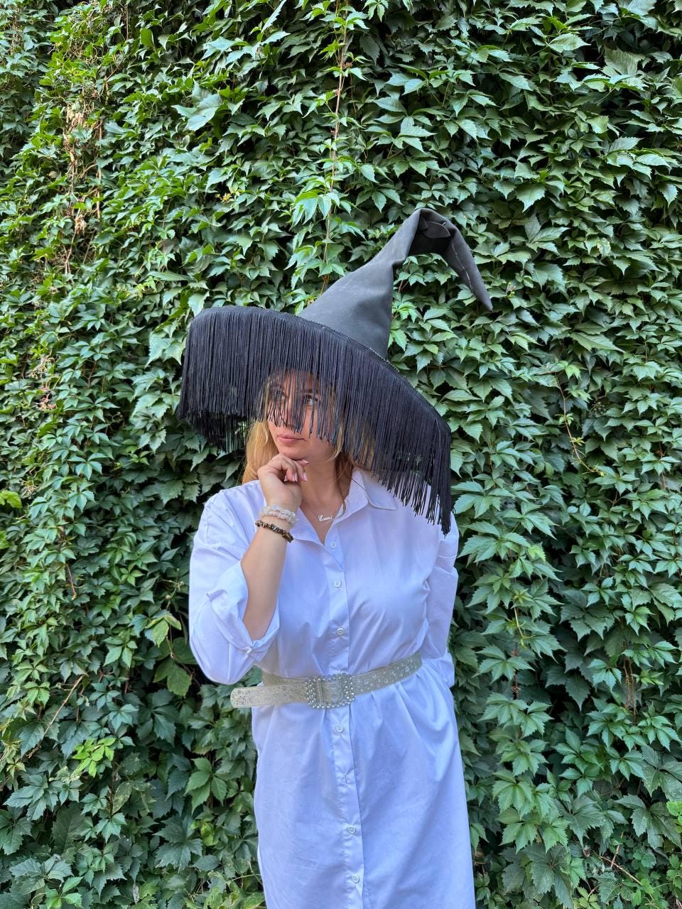 May include: A black witch hat with long fringe. The hat is worn with a white button-down dress and a decorative belt. The background is a wall of green leaves.