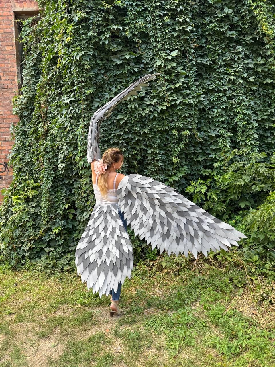 May include: A person wearing large, gray and white feather wings, standing in front of a wall of green ivy. The wings are made of layered, pointed feathers, with a gradient from light to dark gray. The person is wearing a white top and blue jeans.