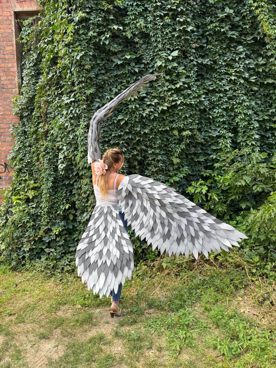 May include: A person wearing large, gray and white feather wings, standing in front of a wall of green ivy. The wings are designed to mimic the appearance of a bird's wings, with layered feathers. The person is wearing a white top and blue jeans.