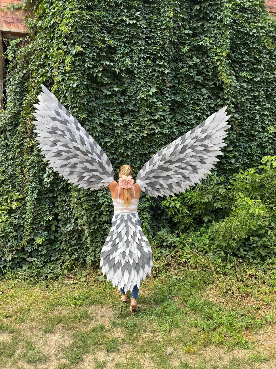 May include: A person wearing a white top, blue jeans, and a skirt and wings made of gray and white feathers. The person is standing in front of a wall of green foliage, with their back to the camera. A pink flower is in their hair.