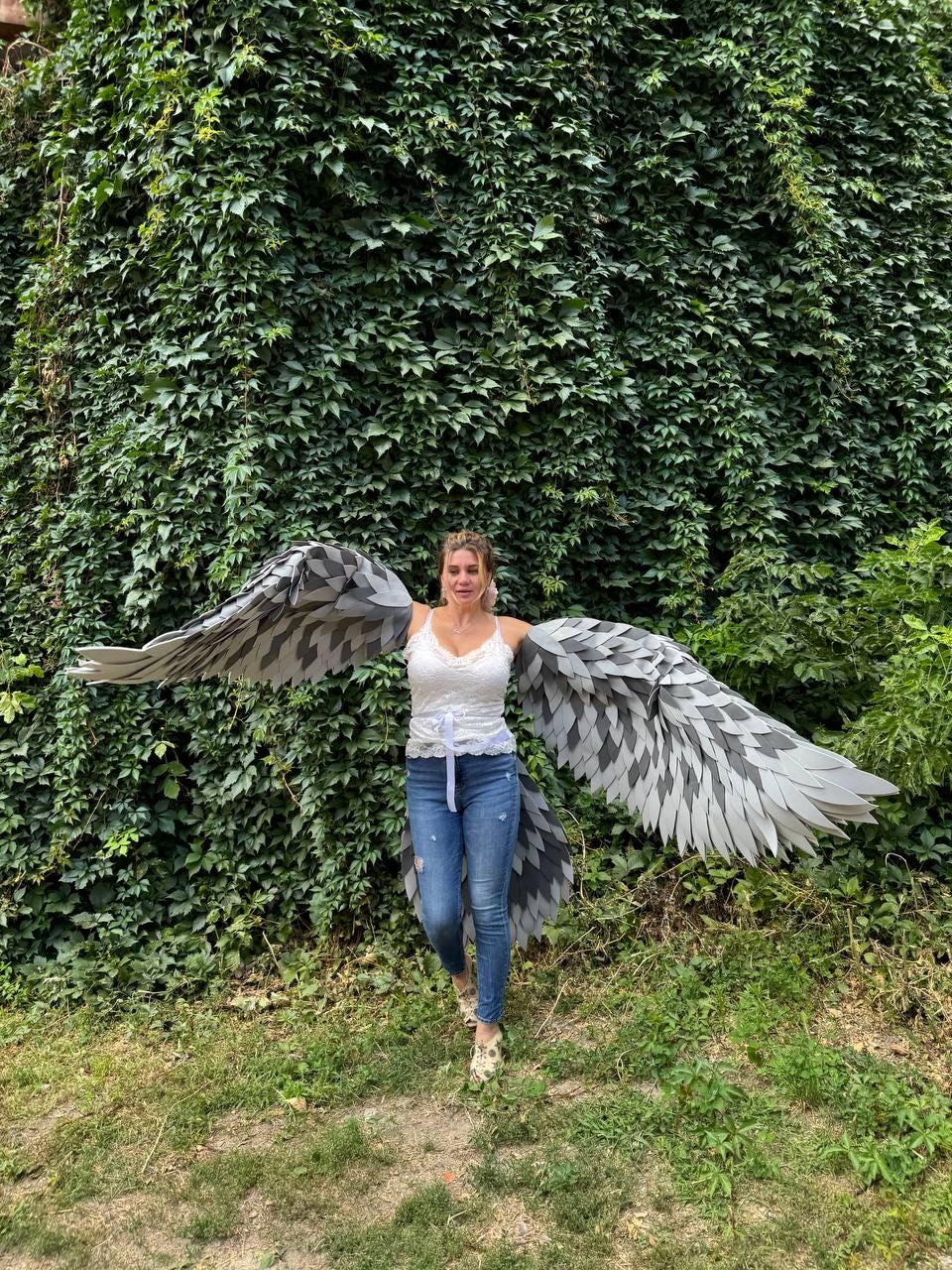 May include: A person wearing large, gray and white feathered wings, standing in front of a wall of green foliage. The wings have a detailed pattern and are spread wide. The person is wearing a white top and blue jeans.
