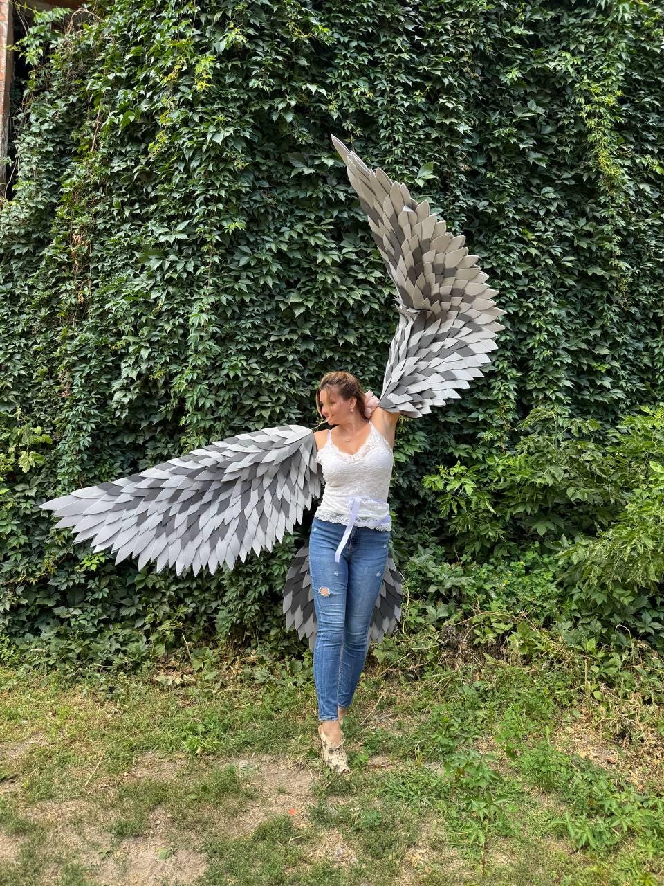 May include: A person wearing large, gray and white feathered wings. The wings are made of overlapping, geometric shapes. The person is wearing a white top and blue jeans, standing in front of a wall of green foliage.