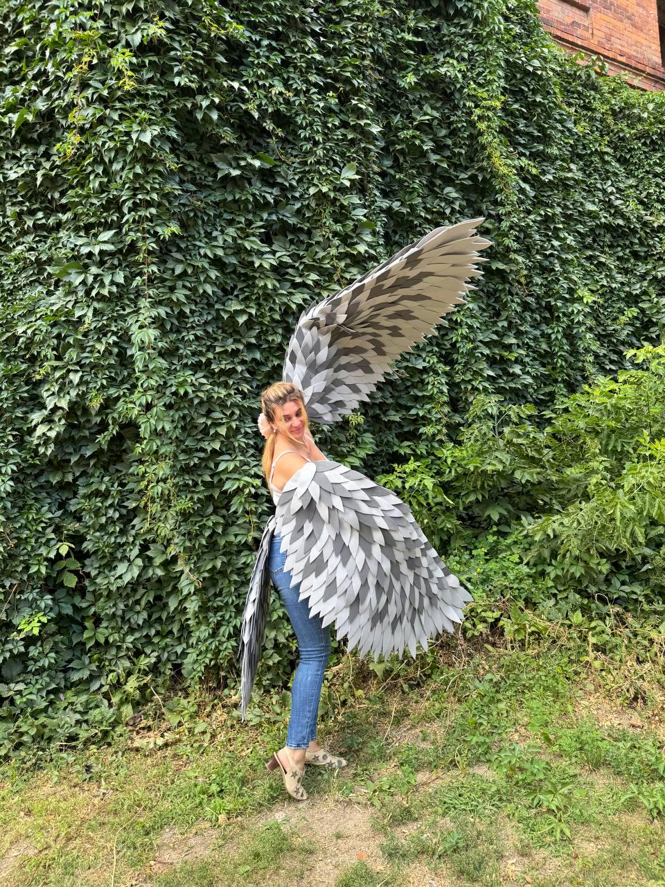 May include: A person wearing large, gray and white feathered wings, posing outdoors. The wings have a layered design with varying shades of gray and white. The person is wearing blue jeans and a white top, standing in front of a wall of green foliage.