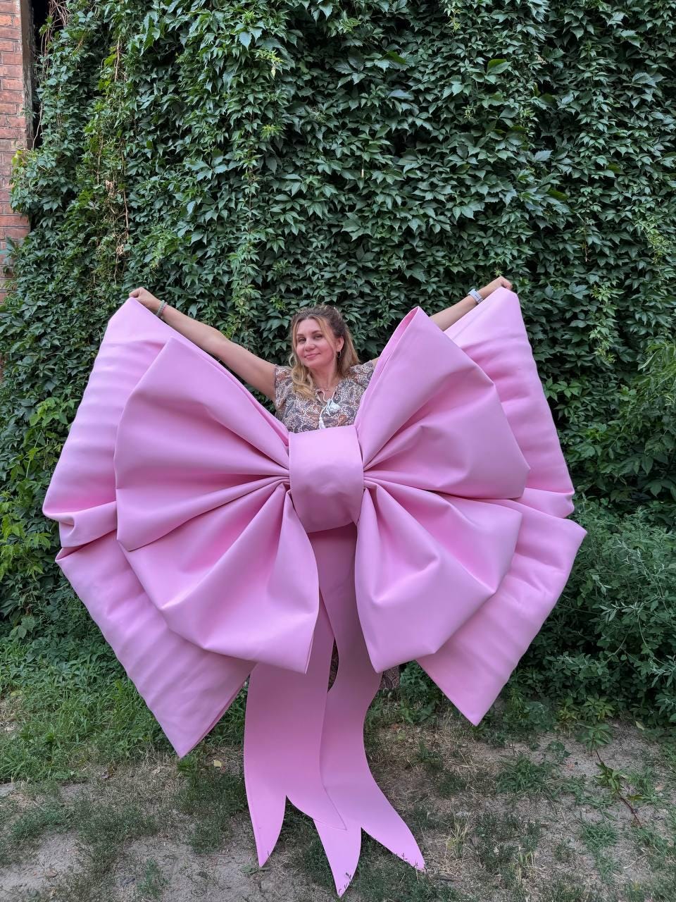 May include: A large, pink bow with long, flowing ribbons. The bow is made of a smooth, solid material and is held up in front of a green leafy background. The bow is a vibrant shade of pink.
