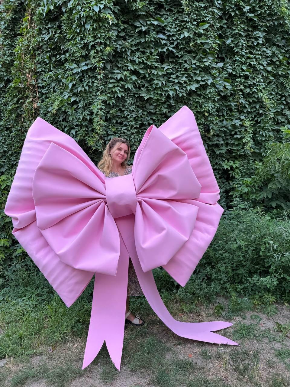May include: A large, pink bow with long, flowing tails. The bow is made of a smooth, possibly satin-like material. The bow is positioned in front of a green leafy background. The bow is large enough to be a prop or decoration.
