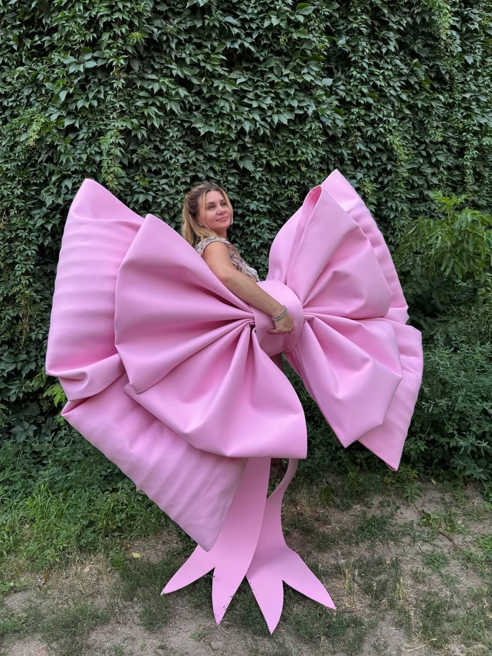 May include: A large, pink bow with a voluminous design. The bow is made of a smooth material and features multiple layers and long, flowing tails. The bow is a vibrant shade of pink and is set against a backdrop of green foliage.
