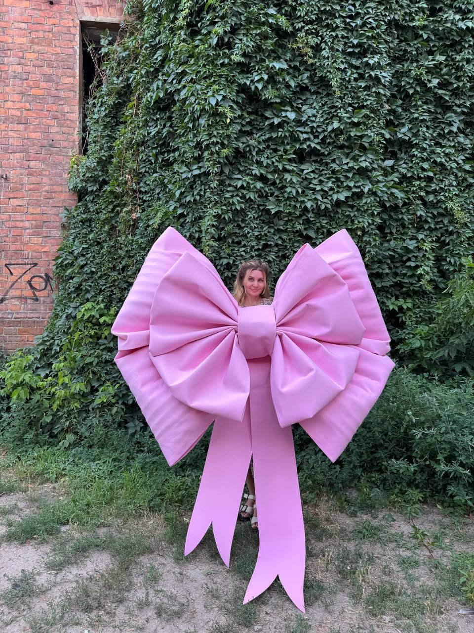 May include: A large, pink bow with long, flowing tails. The bow is made of a smooth, possibly satin-like material. The bow is in front of a wall covered in green vines.