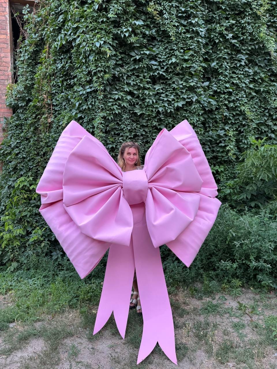 May include: A large, pink bow with long, pointed streamers. The bow is made of a smooth, possibly satin-like material. The bow is large enough to be worn or used as a decorative element. The background features lush green foliage.