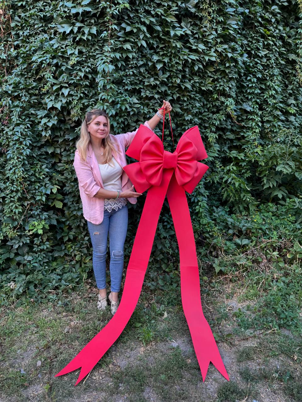 May include: A large, vibrant red bow with long, flowing tails. The bow is held up in front of a lush green wall of foliage. The bow's design features a prominent central knot and wide, elegant streamers, perfect for festive decorations.