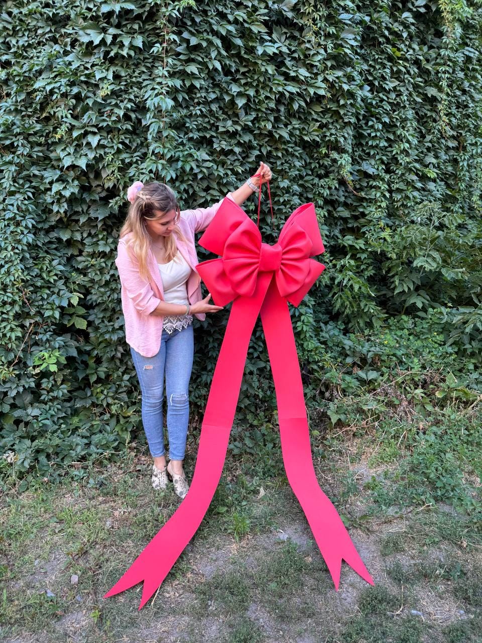 May include: A large, bright red bow with long, flowing ribbons. The bow is held up against a backdrop of green foliage. The bow is a festive decoration, perfect for gifts or events.