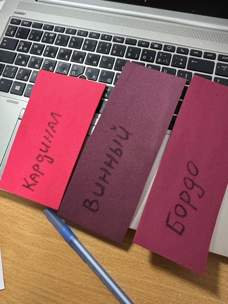 May include: Three rectangular cards in shades of red and purple are displayed on a laptop keyboard. The cards have handwritten text in black. A blue pen lies across the bottom of the cards. The text on the cards is "Кардинал", "Винный", and "Бордо".