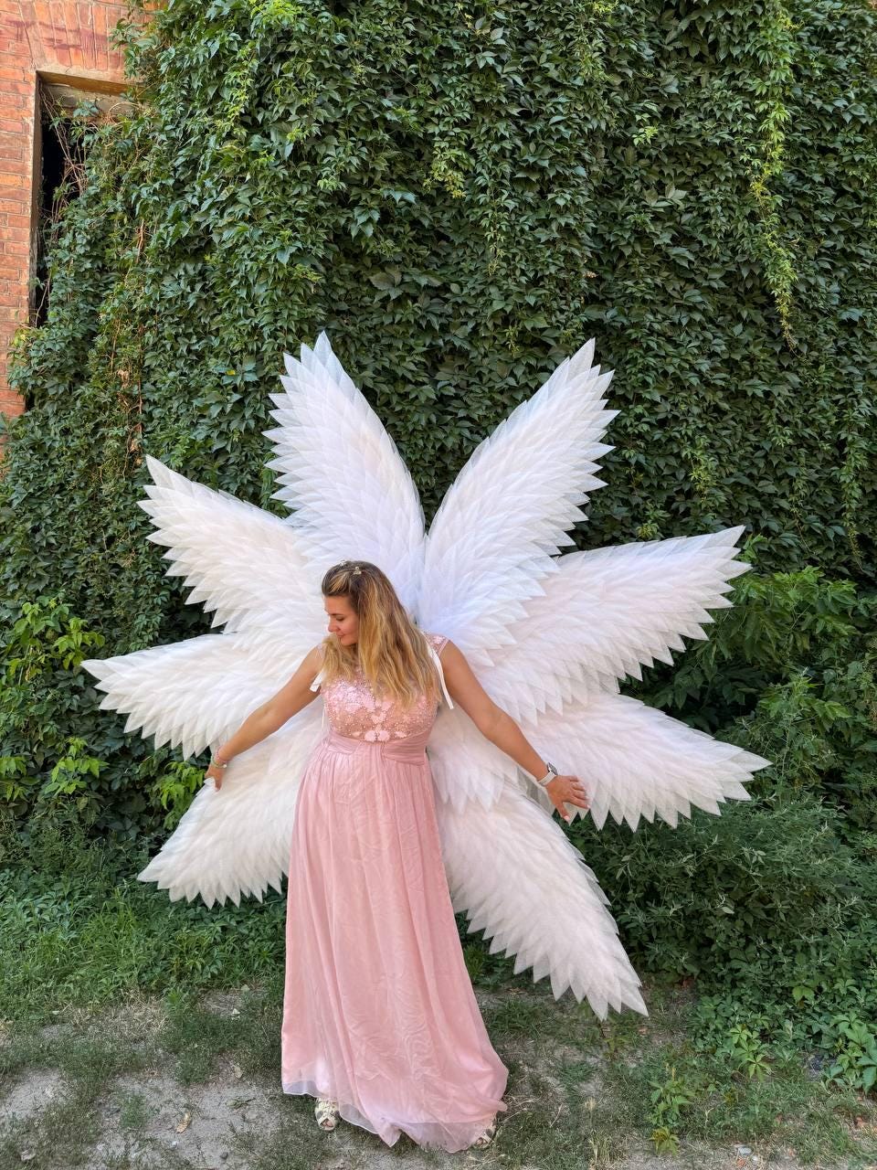 May include: A woman wearing a pink dress and large white angel wings stands in front of a wall of green foliage. The wings are made of many white feathers and are spread out behind her. The dress is a light pink color.