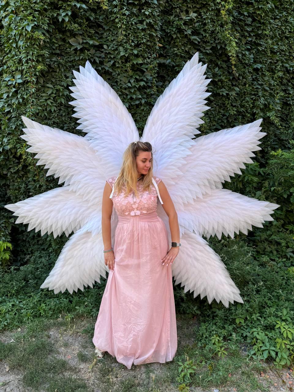 May include: A woman wearing a pink dress and large white angel wings stands in front of a wall of green foliage. The wings are made of white feathers and are attached to the back of the dress. The dress is a light pink color and has a floral design on the bodice.
