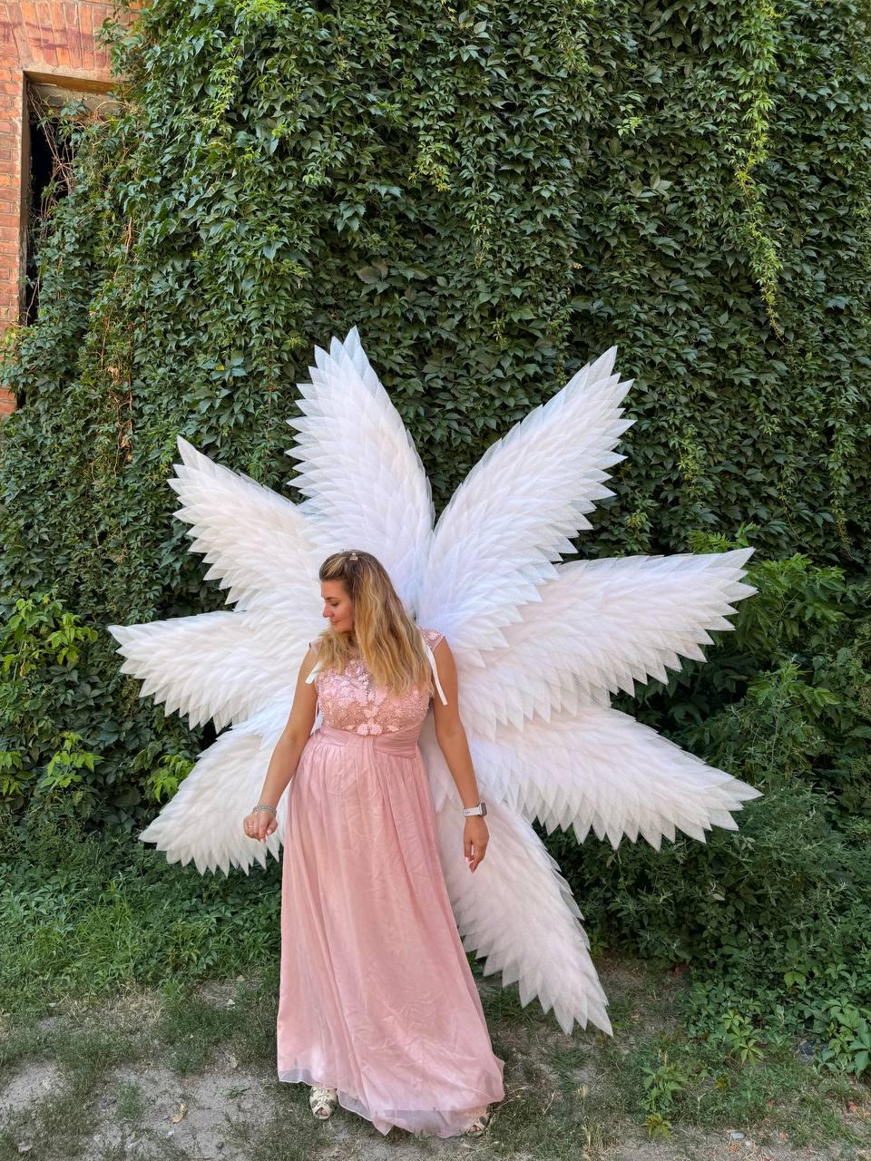 May include: A woman wearing a long, light pink dress and large white angel wings. The wings are made of layered, feathery material. The background features lush green foliage and a brick building.
