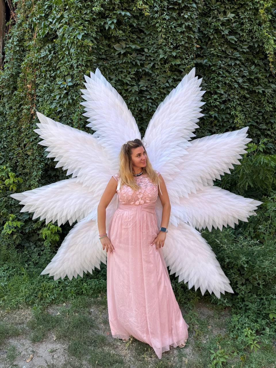 May include: A woman wearing a pink dress and large white angel wings stands in front of a green ivy wall. The dress has a floral bodice and a flowing skirt. The wings are large and detailed.