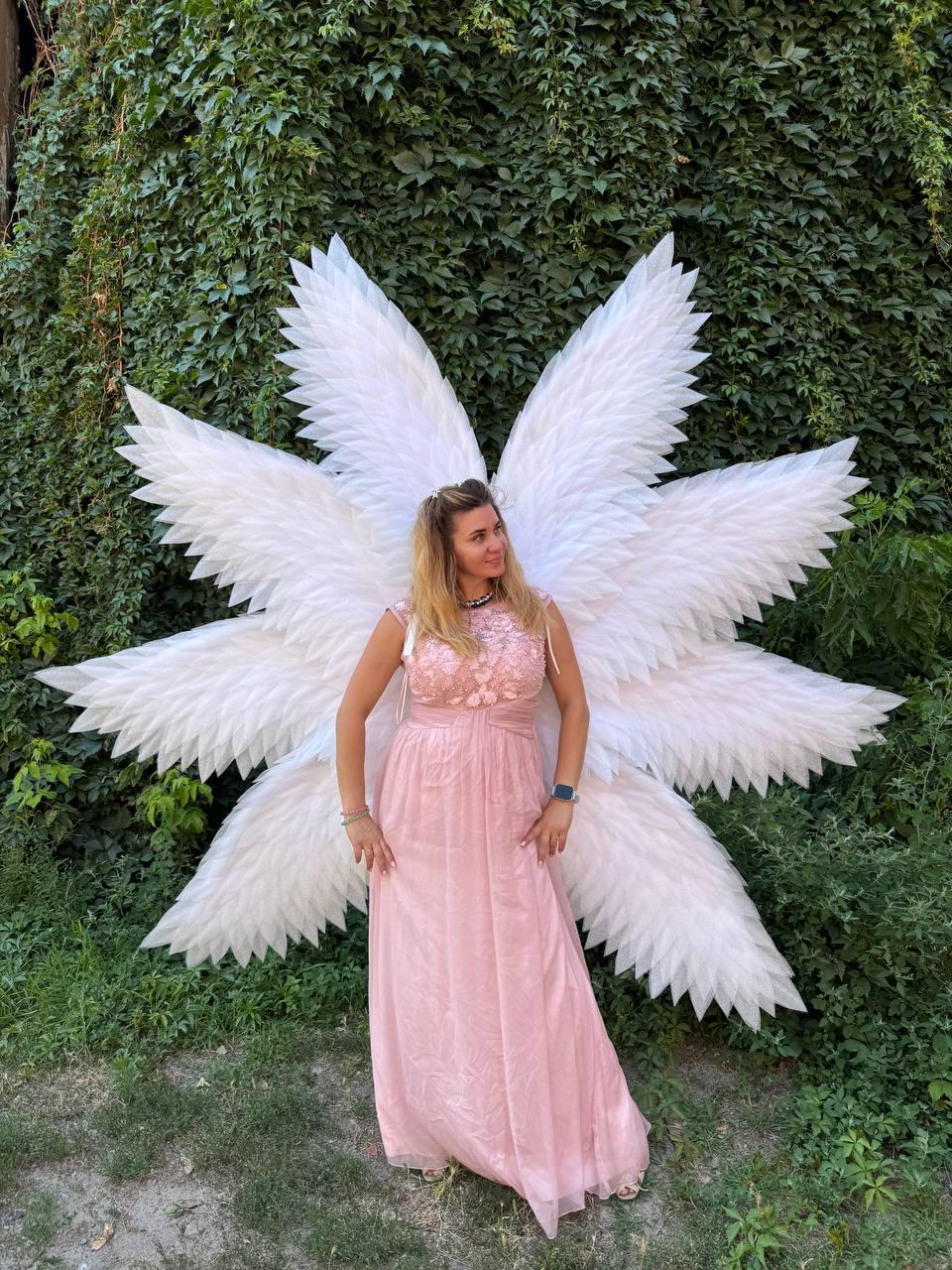May include: A woman wearing a pink dress and large white angel wings stands in front of a green ivy wall. The dress has a floral bodice and a flowing skirt. The wings are large and feathered.