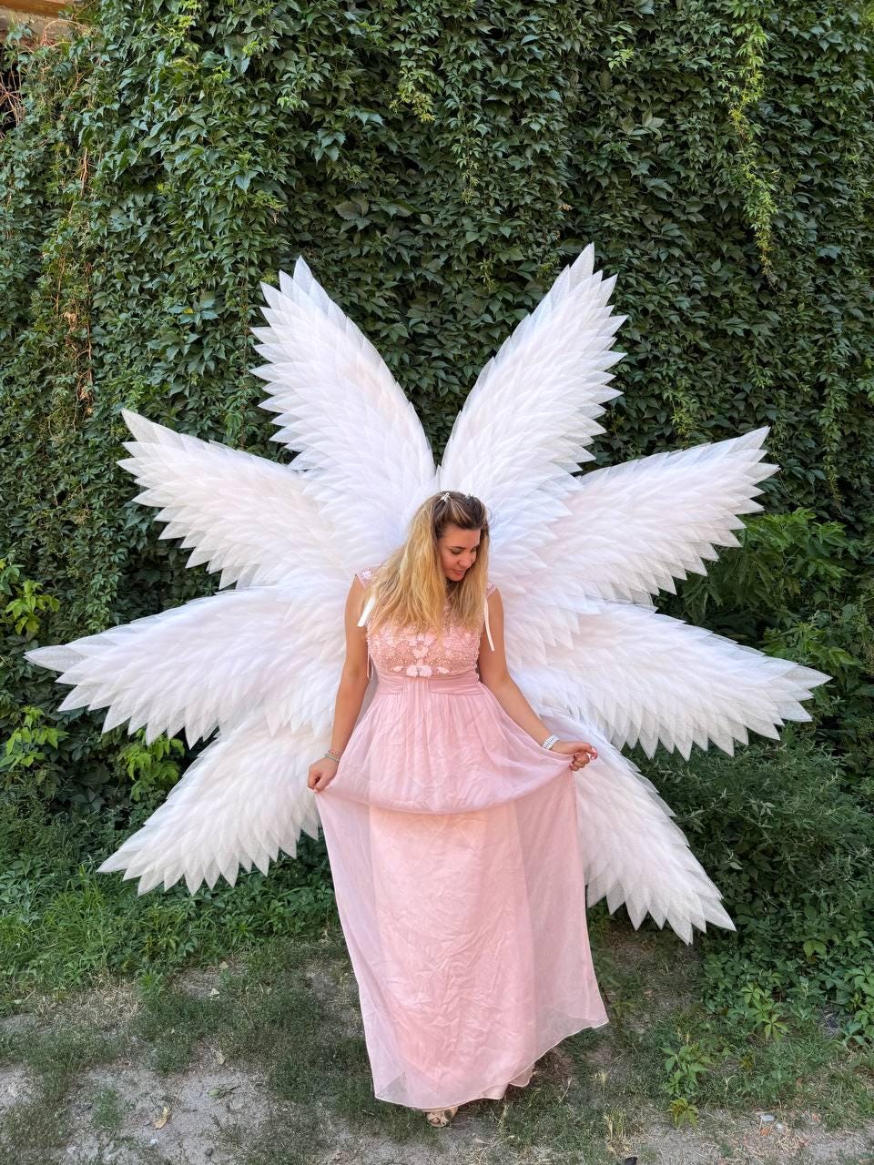 May include: A woman wearing a pink dress and large white angel wings stands in front of a green ivy wall. The wings are large and feathery, with a starburst design. The dress is a light pink color and has a floral bodice.