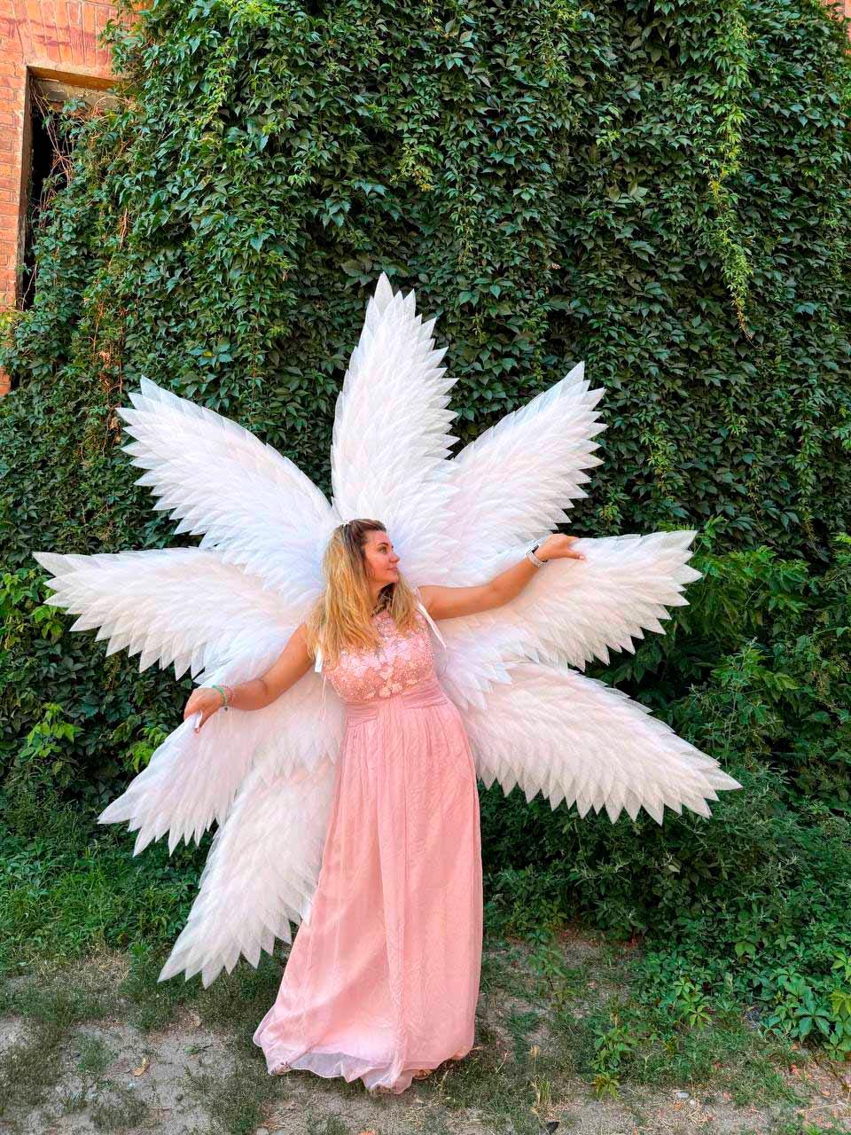 May include: A woman wearing a long, flowing pink dress and large white feathered angel wings stands in front of a wall covered in green ivy. The wings are a prominent feature, with multiple layers of white feathers.