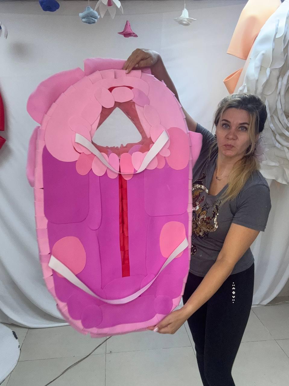 May include: A large, pink and magenta costume piece with a central opening and white straps. The costume has a layered design with rounded edges and a vertical red stripe down the center. Two large pink circles are on the front.