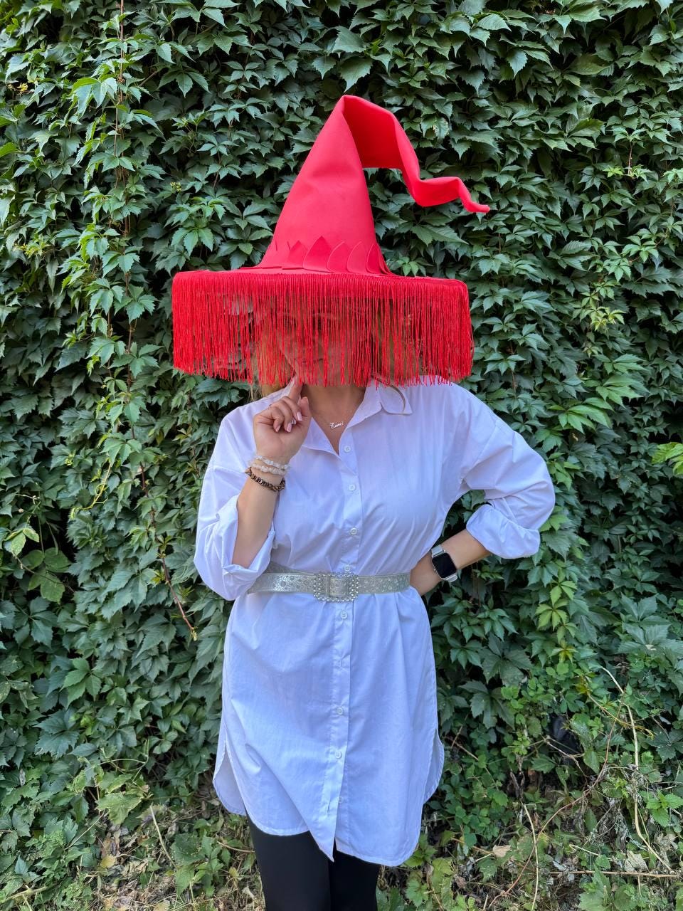 May include: A red witch hat with a wide brim and long fringe. The hat is worn with a white button-down shirt and a silver belt. The background is a wall of green leaves.