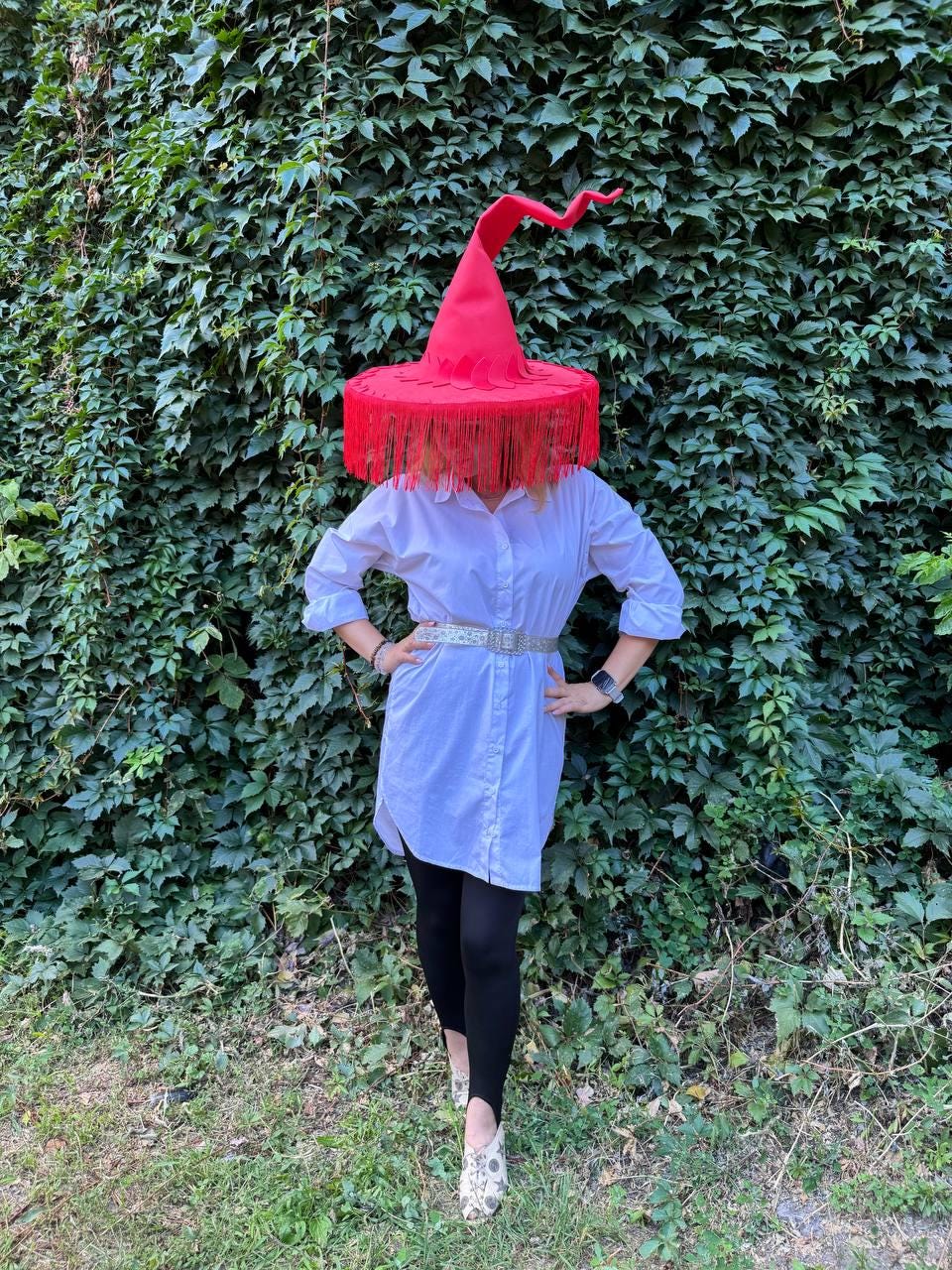 May include: A red witch hat with a wide brim and fringe, worn over a white shirt dress and black leggings. The hat has a tall, curved point. The outfit is accessorized with a silver belt and patterned shoes. The background is a wall of green leaves.