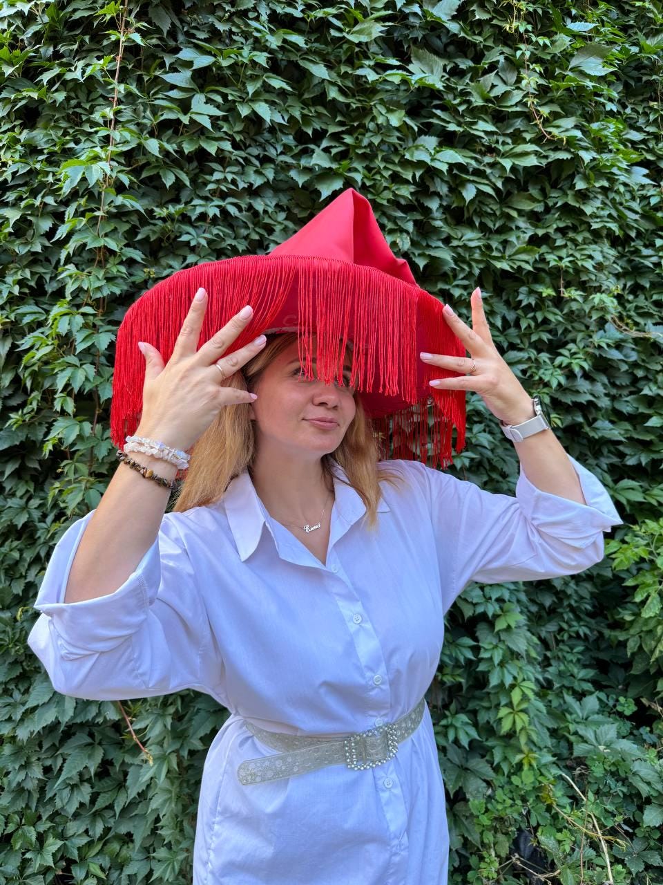 May include: A vibrant red hat with long fringe, paired with a white button-down shirt and a decorative belt. The hat's unique design and bold color make it a statement piece. The background is a lush green wall of leaves.