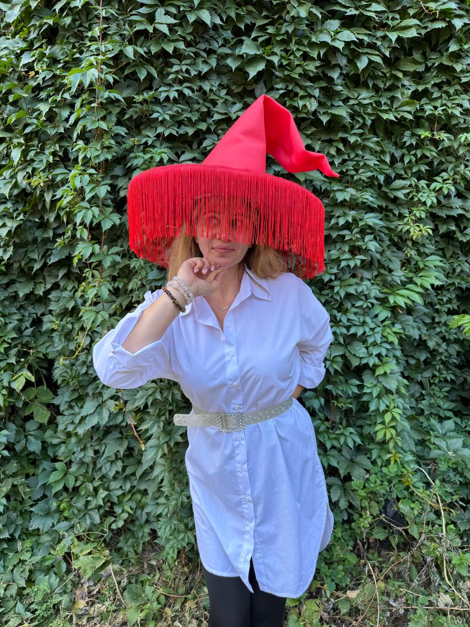 May include: A bright red witch hat with long fringe, worn with a white button-down shirt and a sparkly belt. The hat's pointed crown and fringed brim create a dramatic look. The background is a wall of green leaves.