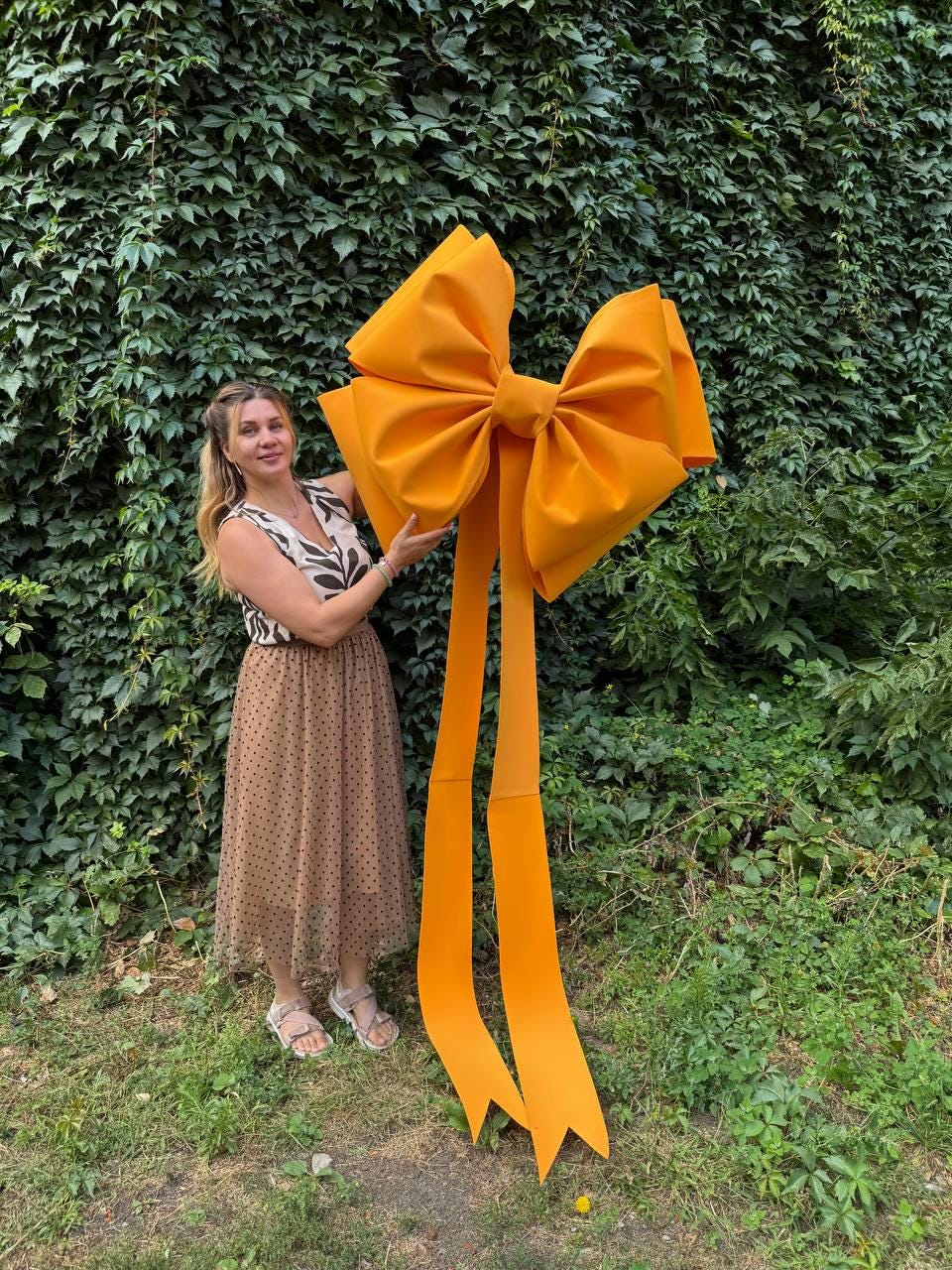 May include: A large, bright orange bow with long, flowing ribbons. The bow is made of a soft material and is positioned in front of a lush green wall of foliage. The bow is a decorative element, suitable for events or celebrations.