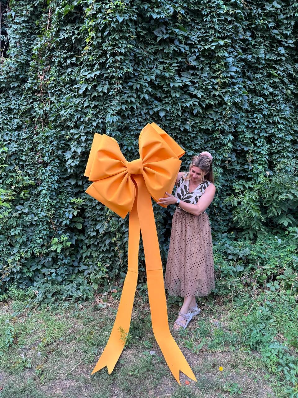 May include: A large, bright orange bow with long, flowing tails. The bow is made of a sturdy material and is positioned in front of a wall of green foliage. The bow is a decorative element, suitable for events or celebrations.
