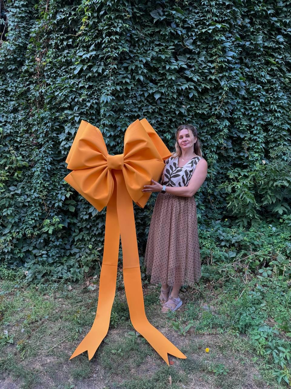 May include: A large, bright orange bow with long, flowing tails. The bow is held in front of a background of green foliage. The bow's size suggests it could be used for a decorative event or as a large-scale decoration.