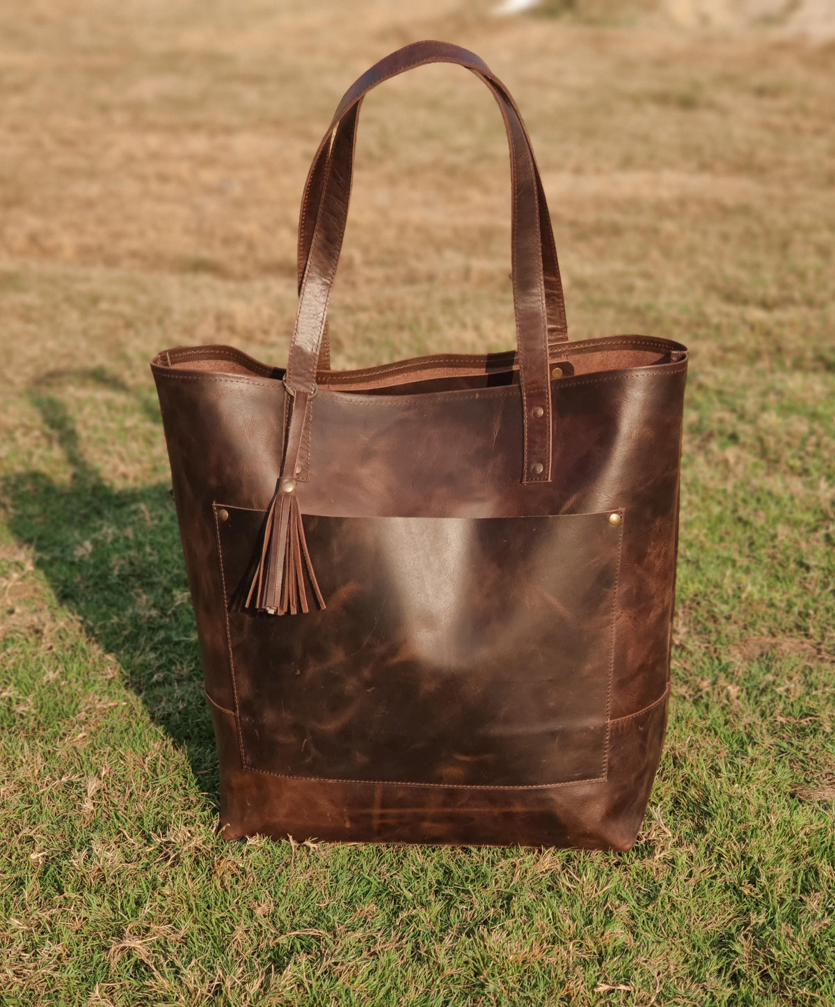 May include: A brown leather tote bag with a front pocket and a tassel. The bag has two handles and is sitting on a grassy surface. The leather has a distressed look.