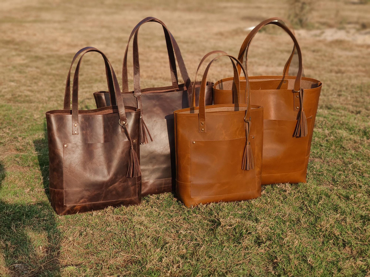 May include: Four leather tote bags in varying shades of brown stand on grass. Each bag has a rectangular shape, top handles, and a decorative tassel. The bags range in color from dark brown to a lighter tan.