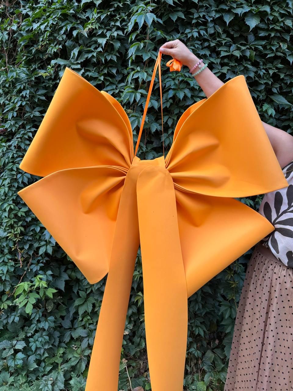 May include: Large, bright orange bow with long streamers, held against a backdrop of green foliage. The bow is made of a thick, matte material, and the streamers hang down. The bow is a vibrant accent piece.