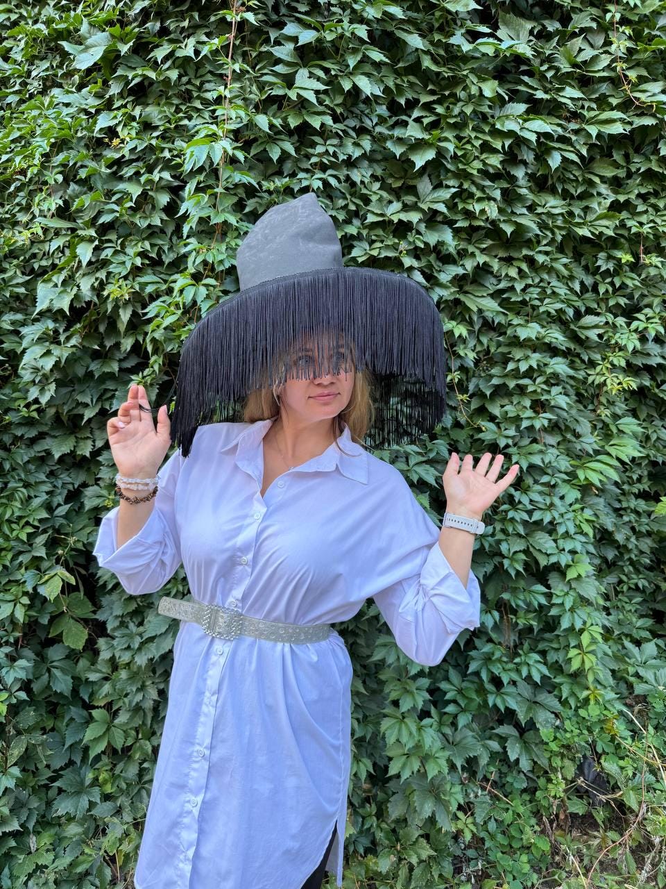 May include: A large, gray hat with a wide brim and long black fringe. The hat is the focal point, set against a backdrop of green foliage. The hat is worn with a white button-down shirt and a silver belt.