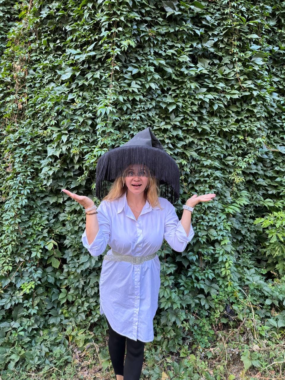 May include: A black witch hat with long fringe is worn by a person in a white button-down shirt and black leggings. The person is standing in front of a wall of green leaves, with arms outstretched.