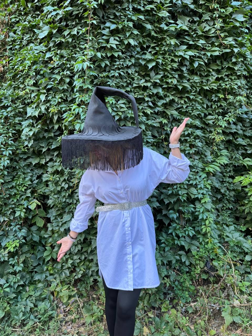 May include: A black witch hat with a wide brim and long fringe. The hat is worn over a white button-down shirt and black leggings. The background is a wall of green leaves.