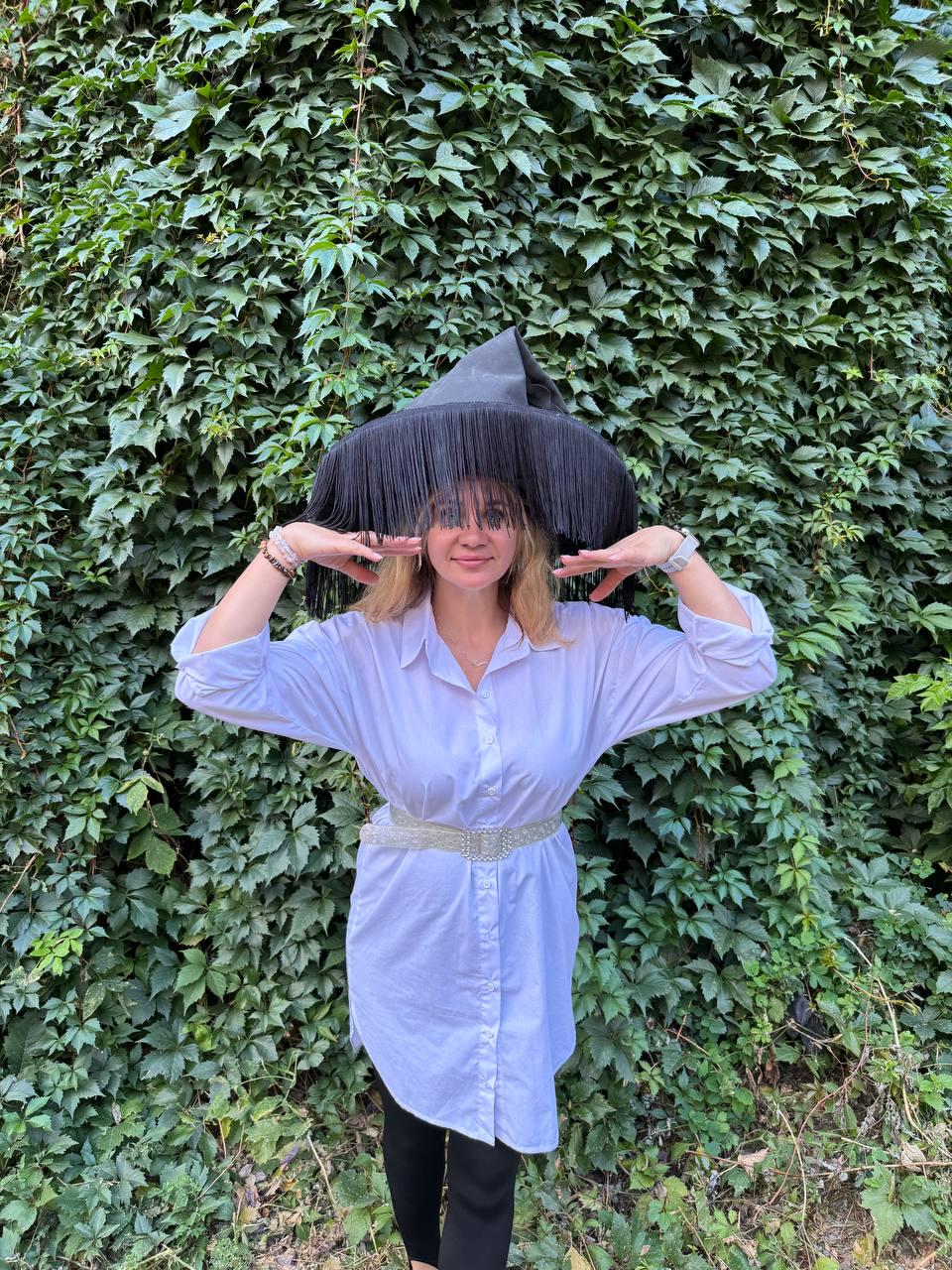 May include: A black witch hat with long fringe is worn over a white button-down shirt and black leggings. The hat has a wide brim and a pointed crown. A silver belt with rhinestones cinches the waist. The background is a wall of green leaves.
