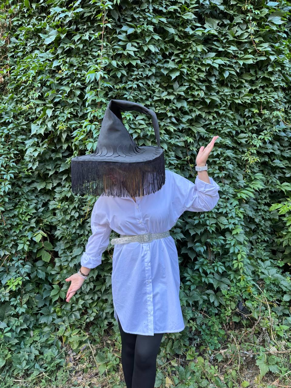 May include: A black witch hat with a wide brim and long fringe. The hat is worn over a white button-down shirt and black leggings. The background is a wall of green leaves.